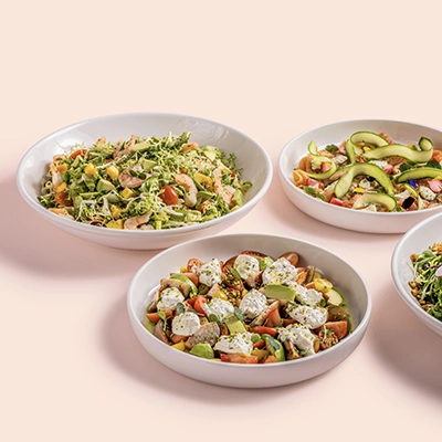 Four colourful salads in white bowls, including avocado, tomato and burrata, on a soft peach background