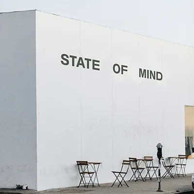 White building wall with the words 'State of Mind' painted in large black letters above café chairs